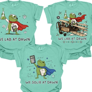 May include: Three light green t-shirts featuring frog illustrations. One shows a frog in a lab coat and red cape holding a beaker with the text WE LAB AT DAWN. Another shows a frog in a plane with the same text. The third shows a frog with a calculator and blue cape with the text WE SOLVE AT DAWN.