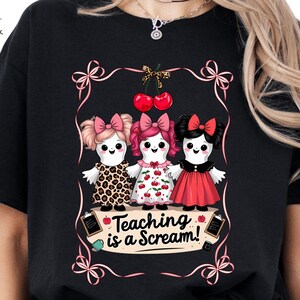 May include: Black t-shirt featuring a whimsical design of three doll-like figures with bows and dresses. The dolls are framed by a pink ribbon border and cherries. The text "Teaching is a Scream!" is displayed on a banner below the dolls.