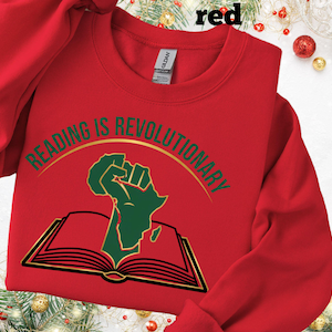 May include: A red crew neck sweatshirt featuring the text "READING IS REVOLUTIONARY" in gold and green. The design includes a green fist and the outline of Africa above an open book. The sweatshirt is a casual style.