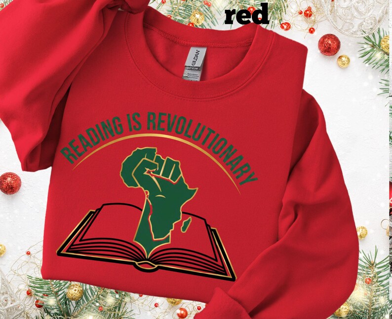 May include: A red crew neck sweatshirt featuring the text "READING IS REVOLUTIONARY" in gold and green. The design includes a green fist and the outline of Africa above an open book. The sweatshirt is a casual style.