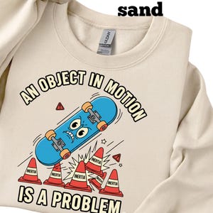 Può includere: Felpa color sabbia con una grafica di uno skateboard blu con una faccia, coni stradali rossi e la scritta "AN OBJECT IN MOTION IS A PROBLEM."