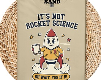 May include: Sand-colored t-shirt with the text "IT'S NOT ROCKET SCIENCE" above a cartoon rocket character holding a clipboard. The rocket is wearing a yellow jacket with a red "R" on it. The text "OH WAIT, YES IT IS" is below the rocket, with star graphics.