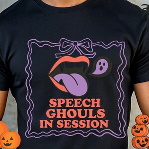 Può includere: T-shirt nera con grafica a tema Halloween. Il design presenta una bocca con zanne, una lingua viola e un fantasma, il tutto all'interno di una cornice viola. Il testo "SPEECH GHOULS IN SESSION" è in arancione e viola.