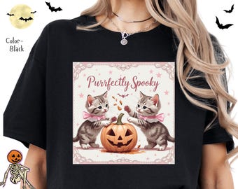 Puede incluir: Camiseta negra con un diseño de Halloween. El diseño presenta dos gatitos grises con lazos rosas, una calabaza tallada y el texto "Purrfectly Spooky" en un marco decorativo. Murciélagos y estrellas completan el diseño.