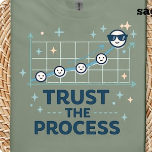 以下が含まれることがあります： セージグリーンのTシャツ。サングラスをかけた笑顔の顔がグラフに描かれており、紺色で「TRUST THE PROCESS」と書かれています。グラフは上昇傾向を示しており、線に沿って他の笑顔の顔があります。