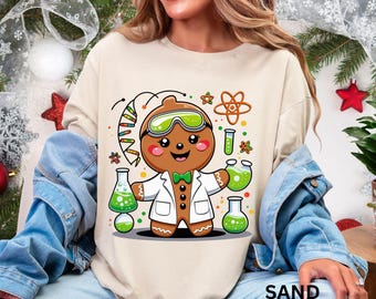 Pode incluir: Camisola cor de areia com um homem de gengibre vestido de cientista. O homem de gengibre usa óculos e um jaleco de laboratório, segurando béqueres com líquido verde. O design inclui gráficos com tema científico e a palavra "SAND" na parte inferior.