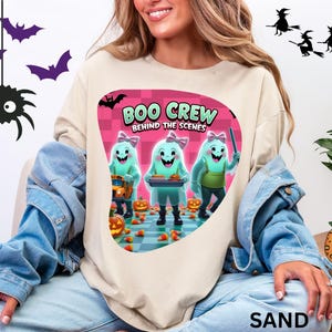 Könnte beinhalten: Sandfarbenes Sweatshirt mit einem Halloween-Motiv. Das Motiv zeigt den Text "BOO CREW BEHIND THE SCENES" und Cartoon-Geister. Das Design ist vor einem rosa karierten Hintergrund mit Kürbissen und Zuckermais platziert.