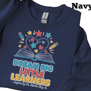 May include: Navy blue sweatshirt with a colorful graphic. The graphic features an open book with a heart-shaped design, stars, pencils, and the text "DREAM BIG LITTLE LEARNERS - Inspired by Dr. Martin King Jr."