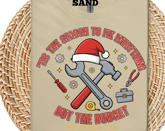 Pode incluir: Camiseta bege com um design natalino. Apresenta ferramentas, um gorro de Papai Noel e o texto "'Tis the Season to Fix Everything But the Budget." Inclui uma chave inglesa, martelo, chave de fenda, alicate, fita métrica e caixa de ferramentas.