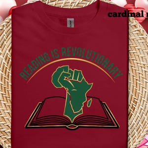 May include: A cardinal red t-shirt with the words "READING IS REVOLUTIONARY" in gold and green. The design includes a green fist and the African continent above an open book.