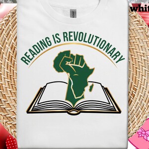 May include: White t-shirt with the text "READING IS REVOLUTIONARY" in green and gold. The design features a green fist rising from an open book, with the outline of the African continent.