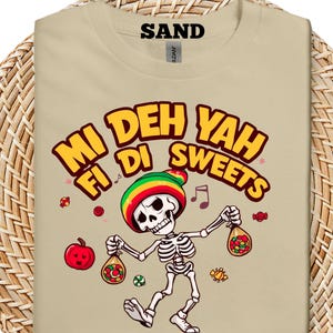 Puede incluir: Camiseta color arena con un esqueleto que lleva un gorro estilo Rasta y sostiene bolsas de caramelos. La camiseta tiene el texto "Mi Deh Yah Fi Di Sweets" en letras amarillas y rojas, con notas musicales y gráficos de caramelos.