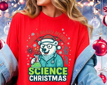 Pode incluir: Uma camisa vermelha de manga comprida com um urso polar usando óculos e um gorro de Papai Noel, segurando um béquer. O texto diz "Have a Beary Merry Science Christmas" e "Science Christmas". Roupa festiva de Natal.