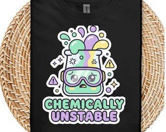 Può includere: T-shirt nera con un disegno di un becher a fumetti con occhiali protettivi, bolle colorate e la scritta "CHEMICALLY UNSTABLE". Il design include simboli di avvertimento e stelle, creando una grafica divertente a tema scientifico.