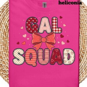 May include: A bright pink t-shirt with the words "GAL SQUAD" in a patchwork design. The letters are decorated with patterns like hearts and stripes, and a large bow sits above the word "SQUAD". The word "heliconia" is printed at the top.