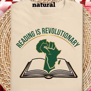 May include: A cream-coloured t-shirt with the slogan "READING IS REVOLUTIONARY" in green and gold. The design shows a green fist in the shape of Africa emerging from an open book. The shirt is made of a soft, comfortable material.