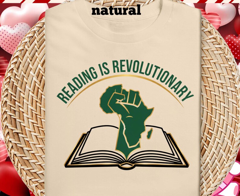May include: A cream-coloured t-shirt with the slogan "READING IS REVOLUTIONARY" in green and gold. The design shows a green fist in the shape of Africa emerging from an open book. The shirt is made of a soft, comfortable material.