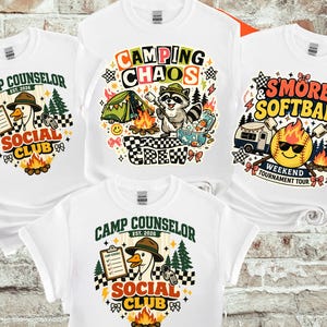 Op de afbeelding: Witte T-shirts met campingthema-ontwerpen. Ontwerpen omvatten een gans met een klembord, een wasbeer die marshmallows roostert en een softbal met een kampvuur. Tekst bevat "Camp Counselor", "Camping Chaos Crew" en "S'mores & Softball."