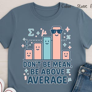 May include: Stone blue t-shirt with a graphic of smiling bar graphs wearing sunglasses. The text reads "DON'T BE MEAN, BE ABOVE AVERAGE" in a playful font. The shirt is paired with blue jeans.
