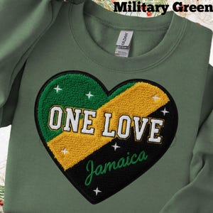 May include: Military green sweatshirt with a heart-shaped design in the colours of the Jamaican flag. The heart features the words "ONE LOVE Jamaica" in white lettering. The sweatshirt is made of a soft material.