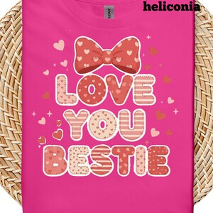 Op de afbeelding: Een felroze t-shirt met de tekst "LOVE YOU BESTIE" in een decoratief lettertype. Het ontwerp bevat een strik, harten en sterren in rode en roze tinten. De merknaam "heliconia" staat bovenaan.
