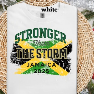 Könnte beinhalten: Weißes T-Shirt mit einem grafischen Design mit dem Text "STRONGER THAN THE STORM" in Grün und Schwarz, darunter "JAMAICA 2025". Das Design enthält die Farben der jamaikanischen Flagge: Grün, Gelb und Schwarz.