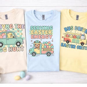 May include: Three pastel-colored t-shirts with Easter-themed graphics. One shirt is cream-colored with a car graphic and the text "Driving the Egg Hunt." Another is light blue with a truck graphic and the text "Serving Easter Energy." The third is yellow with a bus graphic and the text "Bus Driver Easter Crew."