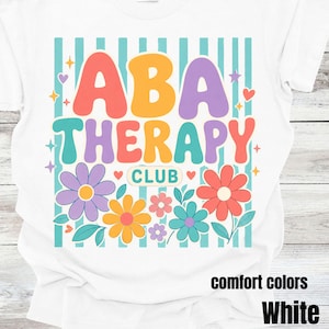 May include: A white t-shirt featuring a graphic design with the text ABA THERAPY CLUB in colorful, rounded letters. The design includes light blue vertical stripes, various flowers in purple, yellow, pink, and red, and small star and heart accents.