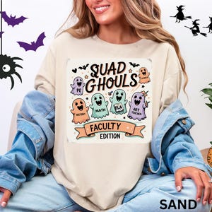 May include: A sand-colored t-shirt with a colorful graphic that reads "Squad Ghouls Faculty Edition." The graphic features cartoon ghosts representing different subjects like PE, History, Math, ELA, Science, and Art Teacher. Halloween-themed decorations are also present.