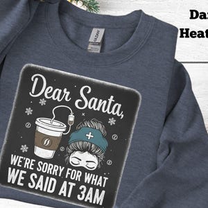 Peut inclure: Sweat-shirt bleu foncé avec l'inscription "Dear Santa, We're sorry for what we said at 3 AM". Le motif comprend une tasse de café, des flocons de neige et une tête d'infirmière avec un bonnet médical. Col rond.