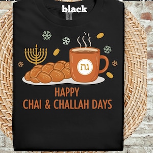 Puede incluir: Camiseta negra con un diseño gráfico que incluye una menorá, pan de jalá y una taza de café. El texto dice "Happy Chai & Challah Days". El diseño se completa con copos de nieve y detalles dorados.