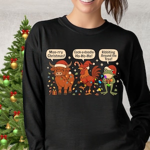May include: Black long-sleeve shirt with a Christmas-themed graphic. The graphic features a cow, rooster, and frog wearing Santa hats and wrapped in Christmas lights. Text includes "Moo-rry Christmas!", "Cock-a-doodle-Ho-Ho-Ho!", and "Ribbiting Around the Tree!"