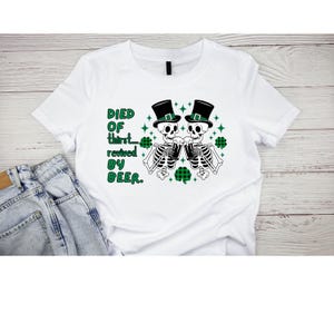 May include: White t-shirt with a green, black, and white graphic of two skeletons wearing green hats and drinking from mugs. The text "DIED OF thirst... revived BY BEER." is printed on the shirt.