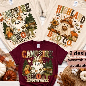 May include: Three t-shirts in cream and maroon featuring graphic prints. Two shirts display a ghost character with a campfire, trees, and the text "CAMPFIRE GHOST SOCIETY". One shirt shows a highland cow in a ghost costume with pumpkins and "HIGHLAND BOO CO".