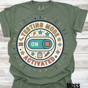 May include: Moss green t-shirt with a circular graphic that reads "TESTING MODE ACTIVATED." The graphic features an "ON" switch, stars, pencils, and other design elements in shades of blue, orange, and yellow. The shirt is a crew neck.