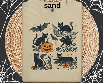 Könnte beinhalten: Sandfarbenes T-Shirt mit einem Halloween-Motiv. Die Grafik zeigt schwarze Katzen, Fledermäuse, Kürbisse und Sterne in Orange und Schwarz. Das Wort "sand" ist oben aufgedruckt. Das T-Shirt ist auf einer gewebten Oberfläche gefaltet.