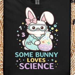 May include: Black t-shirt featuring a cartoon bunny wearing lab goggles and a lab coat, holding a beaker. The shirt has the text "SOME BUNNY LOVES SCIENCE" in colorful letters. The bunny has pink polka dot ears and a pink bow.