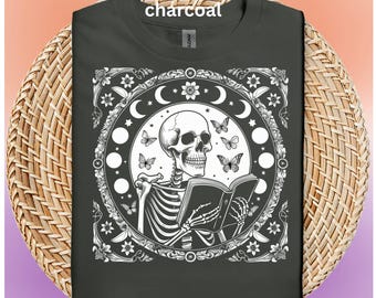May include: Dark grey t-shirt with a white graphic of a skeleton reading a book, encircled by moons, butterflies, and floral patterns. The word "charcoal" is at the top. The shirt is displayed on a woven surface. The design is circular.