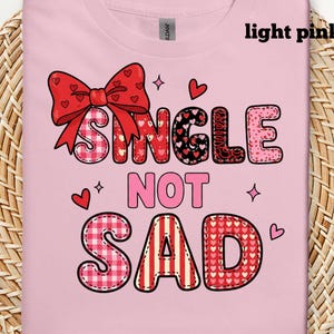 May include: Light pink t-shirt with the text "SINGLE NOT SAD" in a playful font. The design includes a red bow, hearts, and decorative elements. The letters have various patterns, including gingham and stripes.