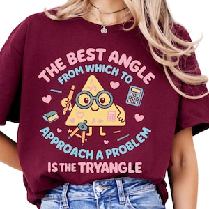 以下が含まれることがあります： メガネをかけた笑顔の三角形が鉛筆を持ち、数学記号に囲まれたグラフィックが特徴のバーガンディ色のTシャツ。「問題を解決するための最良の角度はトライアングルです」と書かれています。