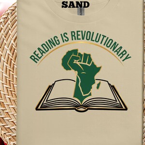 May include: A beige t-shirt with the slogan "READING IS REVOLUTIONARY" in green and gold. The design includes a green fist shaped like Africa above an open book with black outlines. The word "SAND" is printed at the top.