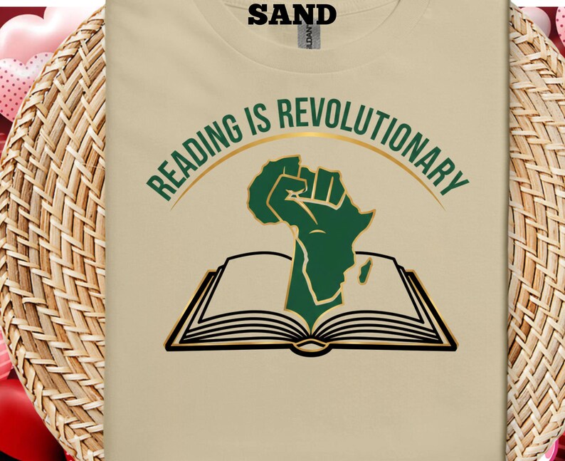 May include: A beige t-shirt with the slogan "READING IS REVOLUTIONARY" in green and gold. The design includes a green fist shaped like Africa above an open book with black outlines. The word "SAND" is printed at the top.