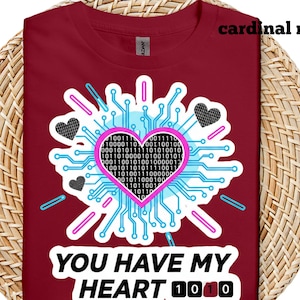 Pode incluir: Camiseta vermelha cardinal com um design gráfico. O design apresenta um coração rosa preenchido com código binário, rodeado por um design de placa de circuito e duas formas de coração pretas. O texto diz "YOU HAVE MY HEART 1010".