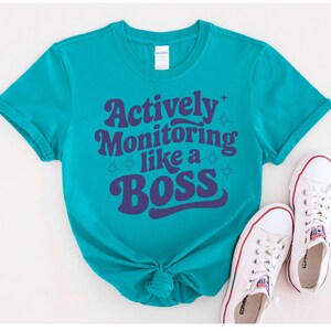 May include: Teal blue short-sleeved t-shirt with a purple graphic that says "Actively Monitoring Like a Boss" in a retro font.