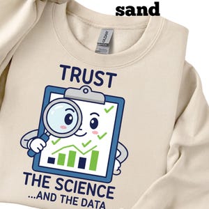 Può includere: Felpa color sabbia con la scritta "TRUST THE SCIENCE...AND THE DATA". La grafica presenta una clip board con una lente d'ingrandimento, grafici e segni di spunta. La felpa è realizzata in un materiale morbido.