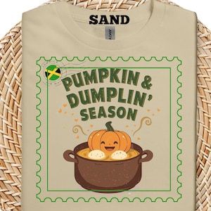 May include: Sand-colored t-shirt featuring a graphic of a pumpkin and dumplings in a pot, with the text "PUMPKIN & DUMPLIN' SEASON" in green. The design is framed with a green border and a Jamaican flag. A fun, food-themed tee.