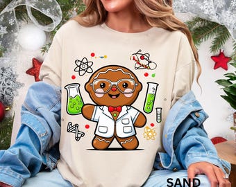 Pode incluir: Camisola cor de areia com um boneco de gengibre de desenho animado vestido de cientista, usando óculos e um jaleco de laboratório. O design inclui béqueres com líquido verde, átomos e a palavra "SAND" na parte inferior.