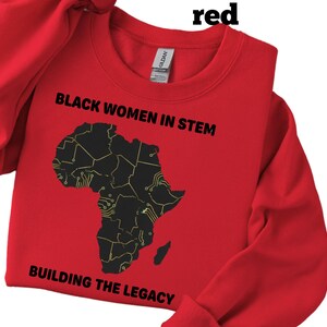 May include: A red sweatshirt featuring the text "BLACK WOMEN IN STEM" and a black graphic of the African continent. The text "BUILDING THE LEGACY" is below the graphic. The sweatshirt is made of a soft material.