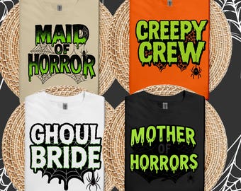 May include: Four Halloween-themed t-shirts with spooky phrases. The shirts are beige, orange, white, and black. Each shirt has a different phrase in green and black, including "Maid of Horror," "Creepy Crew," "Ghoul Bride," and "Mother of Horrors."