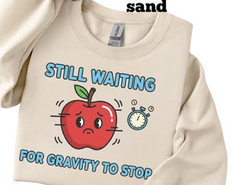 May include: Sand-colored sweatshirt with the text "STILL WAITING FOR GRAVITY TO STOP". The design features a red apple with a sad face and a blue stopwatch. The sweatshirt is made of a soft material.